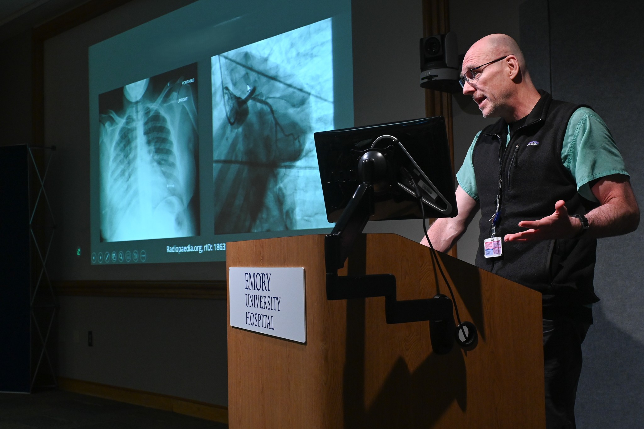 person standing at a wooden podium with the sign Emory Healthcare on it, talking with one arm raised with palm up to chest level and talking
